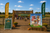 A farm entrance sign reading 'PETTING ZOO' with people and animals walking through the gate, flanked by tall 'Petting Zoo' feather flags and a 'Barnyard Brouhaha!' sign in a sunny outdoor setting.